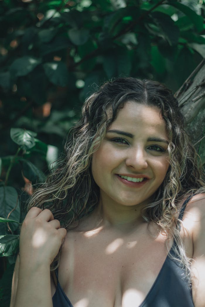 Portrait of a smiling woman outdoors, surrounded by greenery under dappled sunlight.