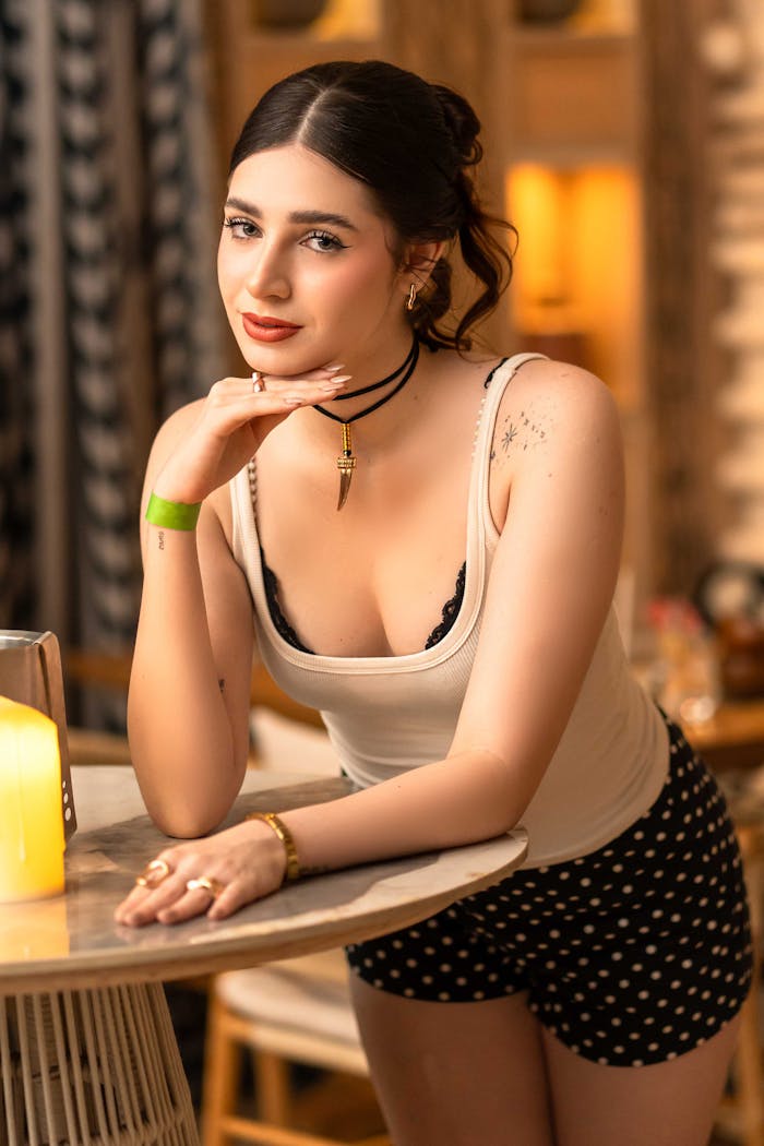 Chic woman in stylish attire leaning on table in ambient indoor setting, exuding elegance and charm.