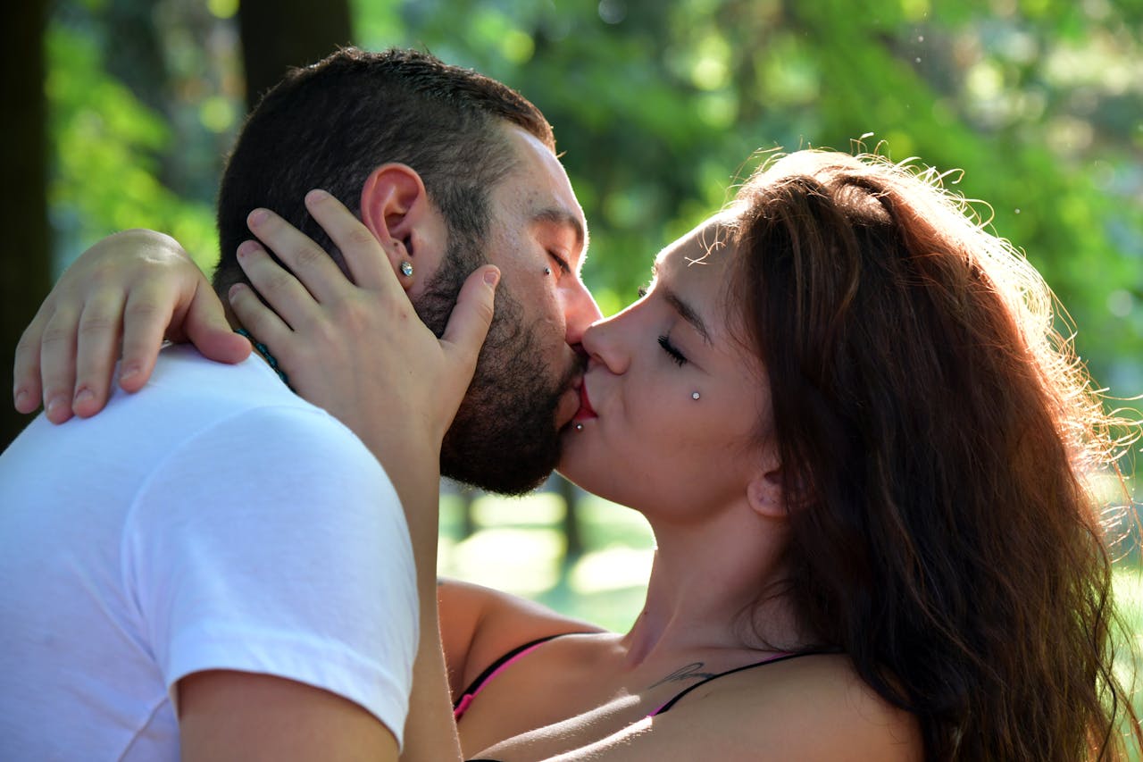 A couple sharing a tender kiss in a sunlit park, surrounded by lush greenery.